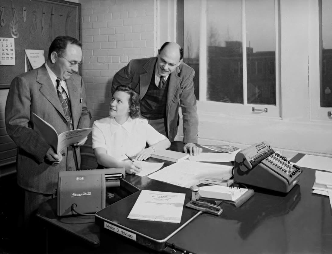 A woman at her desk in 1951 looking up at two colleagues, a calculator beside her and her name on a plate at the desk’s edge